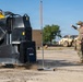 A person in military-style camouflage clothing and a face mask is giving a thumbs-down hand signal near heavy construction equipment on a gravel surface. The scene appears to be a military or construction staging area with multiple pieces of heavy machinery visible. An orange-tipped marker pole is positioned between the worker and the equipment.