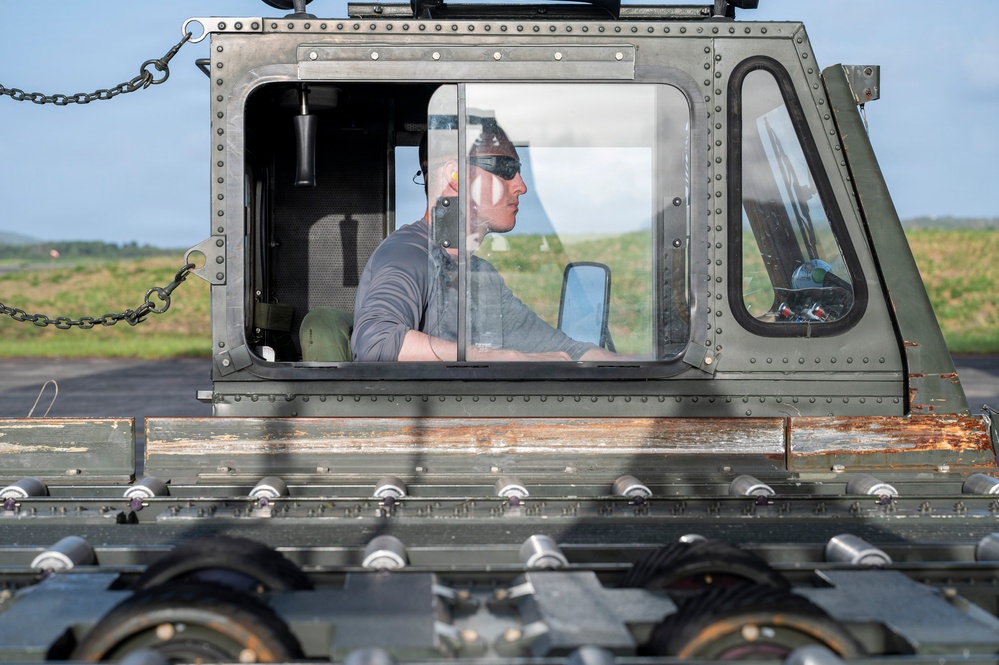 A person is seated inside the enclosed cab of a military ground support vehicle, likely on an airfield or tarmac. The vehicle appears to be an olive-drab painted aircraft arresting system or runway equipment vehicle, featuring a roller/cable assembly visible in the foreground. The operator is wearing sunglasses and appears to be actively operating or monitoring the equipment.