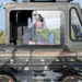 A person is seated inside the enclosed cab of a military ground support vehicle, likely on an airfield or tarmac. The vehicle appears to be an olive-drab painted aircraft arresting system or runway equipment vehicle, featuring a roller/cable assembly visible in the foreground. The operator is wearing sunglasses and appears to be actively operating or monitoring the equipment.