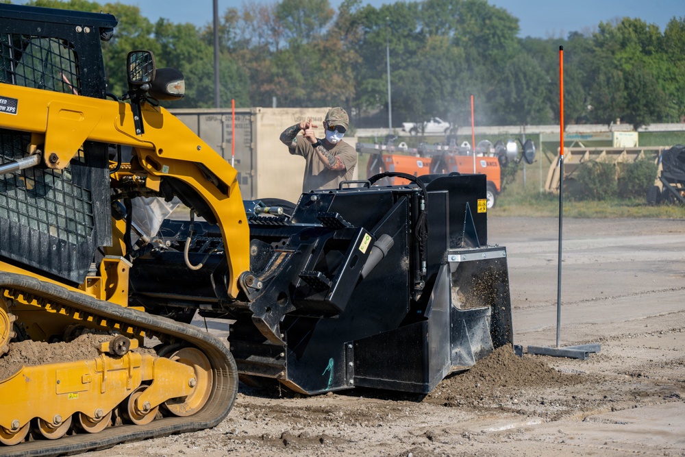 A worker wearing a face mask and cap is operating or inspecting a large black attachment on a yellow tracked skid steer loader at a construction or grading site. The attachment appears to be a soil compactor or pulverizer being used on a dirt surface. The scene takes place in an open outdoor area with trees, orange marker poles, and additional equipment visible in the background.