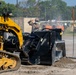 A worker wearing a face mask and cap is operating or inspecting a large black attachment on a yellow tracked skid steer loader at a construction or grading site. The attachment appears to be a soil compactor or pulverizer being used on a dirt surface. The scene takes place in an open outdoor area with trees, orange marker poles, and additional equipment visible in the background.