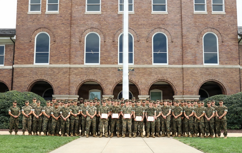 A large group of military personnel in matching camouflage uniforms pose for a formal group photograph in front of a multi-story red brick building with arched windows. Several individuals in the center of the front row are holding certificates or documents, suggesting a graduation, award, or recognition ceremony. A flagpole is visible centered behind the group.