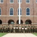 A large group of military personnel in matching camouflage uniforms pose for a formal group photograph in front of a multi-story red brick building with arched windows. Several individuals in the center of the front row are holding certificates or documents, suggesting a graduation, award, or recognition ceremony. A flagpole is visible centered behind the group.
