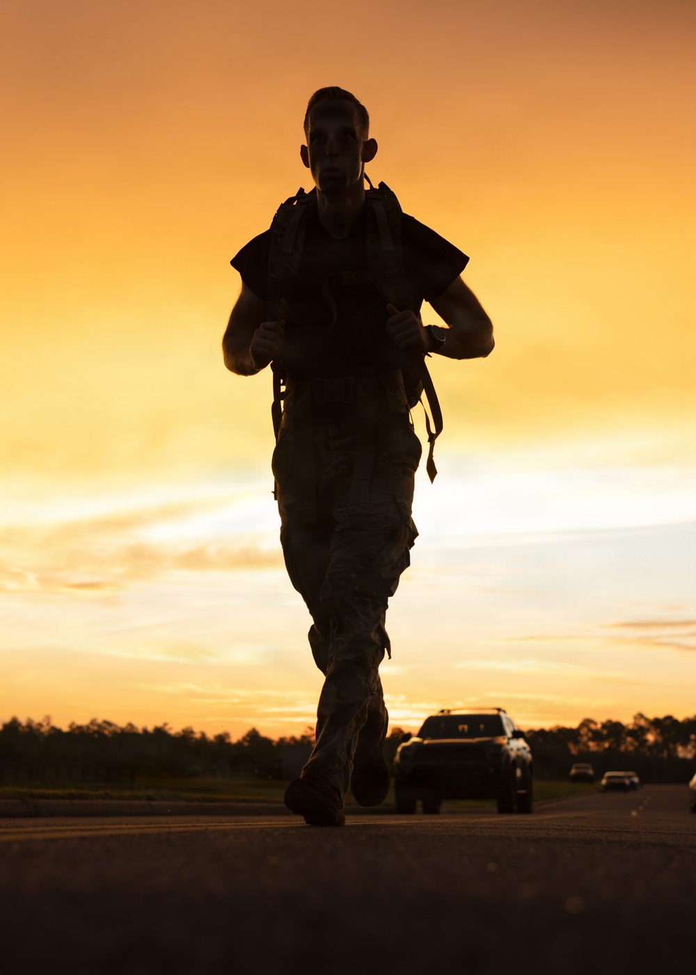 A silhouetted individual in military uniform and carrying a backpack/rucksack is running along a paved road at sunset or sunrise. The warm orange and golden sky creates a dramatic backlit effect, rendering the subject almost entirely in silhouette. Vehicles and a tree line are visible in the background.
