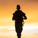 A silhouetted individual in military uniform and carrying a backpack/rucksack is running along a paved road at sunset or sunrise. The warm orange and golden sky creates a dramatic backlit effect, rendering the subject almost entirely in silhouette. Vehicles and a tree line are visible in the background.