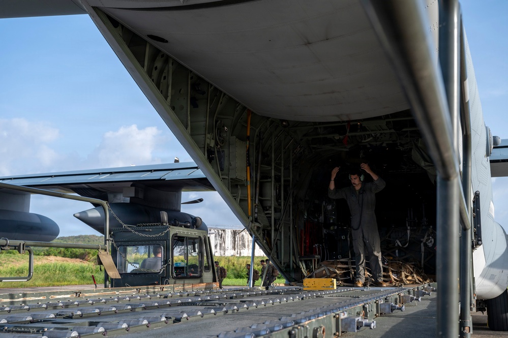 A military loadmaster or crew member stands inside the open rear cargo ramp of a large military transport aircraft, appearing to give hand signals to guide loading operations. A military helicopter, secured with chains, is visible on the cargo ramp and partially inside the aircraft. The scene takes place on an airfield tarmac with green vegetation and partly cloudy skies visible in the background.