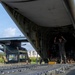 A military loadmaster or crew member stands inside the open rear cargo ramp of a large military transport aircraft, appearing to give hand signals to guide loading operations. A military helicopter, secured with chains, is visible on the cargo ramp and partially inside the aircraft. The scene takes place on an airfield tarmac with green vegetation and partly cloudy skies visible in the background.