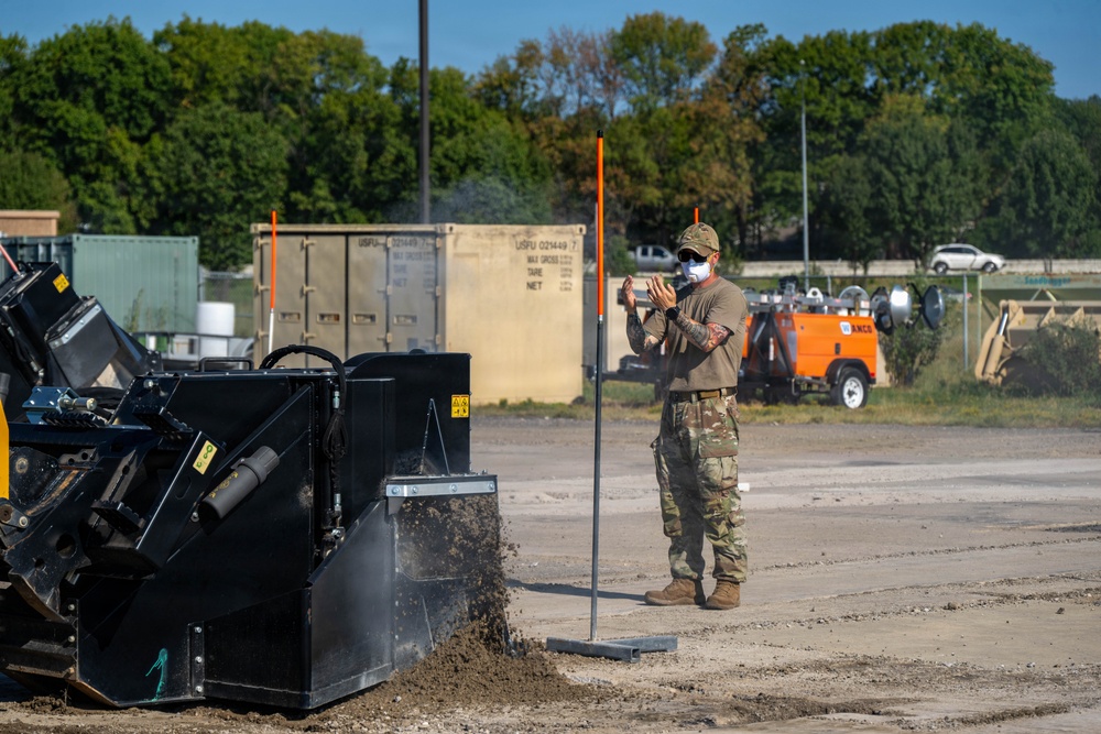 A person in military camouflage uniform and face mask is directing or signaling near heavy construction equipment on what appears to be a military installation or construction site. The individual stands beside an orange survey/marker pole while a skid-steer loader or similar machinery operates nearby, displacing soil. The background shows military shipping containers, an orange utility vehicle, and tree line.