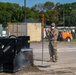 A person in military camouflage uniform and face mask is directing or signaling near heavy construction equipment on what appears to be a military installation or construction site. The individual stands beside an orange survey/marker pole while a skid-steer loader or similar machinery operates nearby, displacing soil. The background shows military shipping containers, an orange utility vehicle, and tree line.