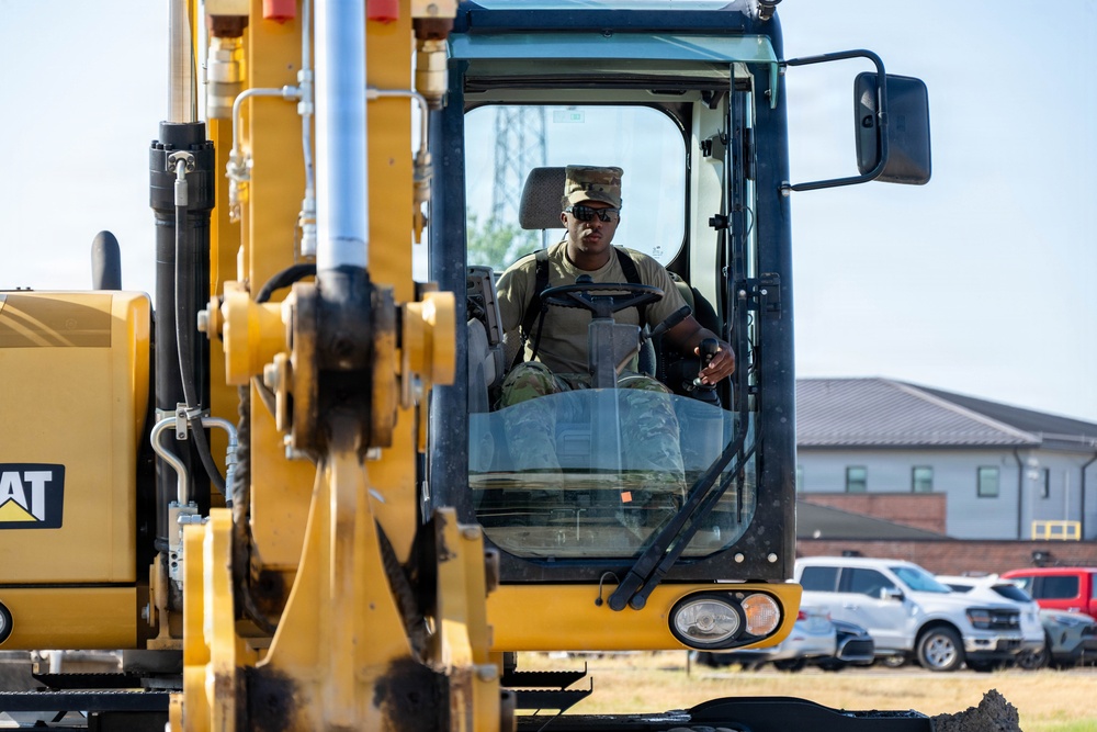 A military service member in camouflage uniform and patrol cap is operating a Caterpillar (CAT) heavy construction machine, likely an excavator or motor grader. The operator is seated in the enclosed cab, wearing sunglasses, and appears to be actively controlling the equipment. The setting appears to be a military installation based on the surrounding buildings and vehicle parking area visible in the background.