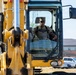 A military service member in camouflage uniform and patrol cap is operating a Caterpillar (CAT) heavy construction machine, likely an excavator or motor grader. The operator is seated in the enclosed cab, wearing sunglasses, and appears to be actively controlling the equipment. The setting appears to be a military installation based on the surrounding buildings and vehicle parking area visible in the background.