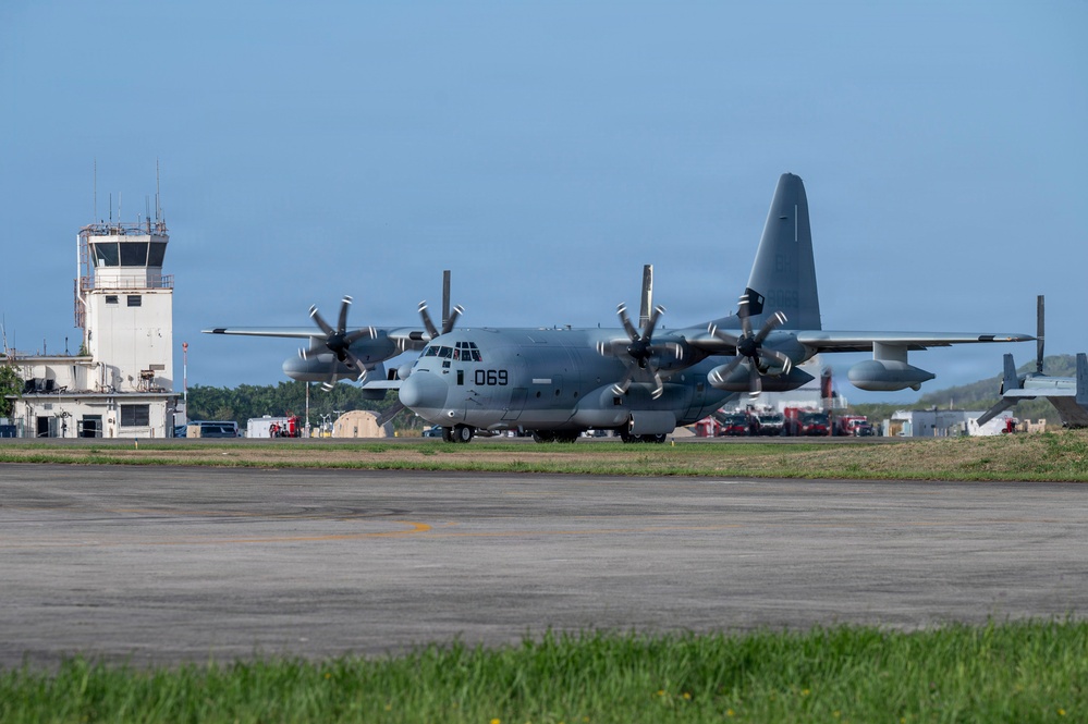A military turboprop transport aircraft marked '069' is taxiing on a runway apron at a military airfield. The aircraft appears to be a C-130J Super Hercules in a gray paint scheme, consistent with U.S. Marine Corps markings. An air traffic control tower and various ground support vehicles are visible in the background.