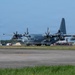 A military turboprop transport aircraft marked '069' is taxiing on a runway apron at a military airfield. The aircraft appears to be a C-130J Super Hercules in a gray paint scheme, consistent with U.S. Marine Corps markings. An air traffic control tower and various ground support vehicles are visible in the background.