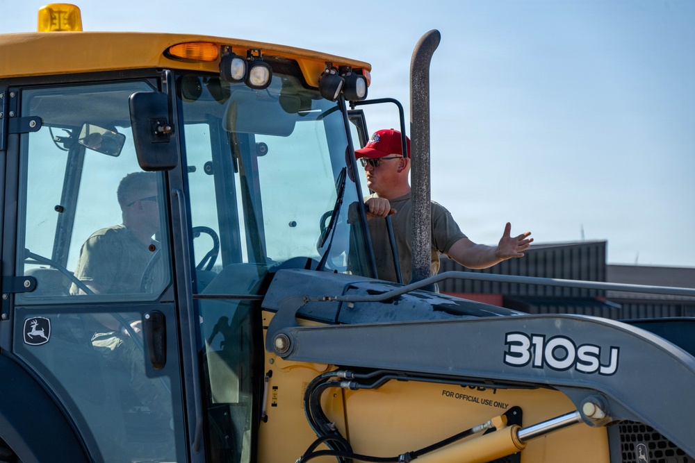 A man wearing a red cap and sunglasses is standing on or beside a John Deere 310SJ backhoe loader, gesturing with his right hand toward the operator seated inside the cab. The machine is yellow and bears a 'FOR OFFICIAL USE ONLY' label, suggesting a military or government context. The scene appears to be an outdoor equipment operation or training exercise.