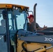 A man wearing a red cap and sunglasses is standing on or beside a John Deere 310SJ backhoe loader, gesturing with his right hand toward the operator seated inside the cab. The machine is yellow and bears a 'FOR OFFICIAL USE ONLY' label, suggesting a military or government context. The scene appears to be an outdoor equipment operation or training exercise.