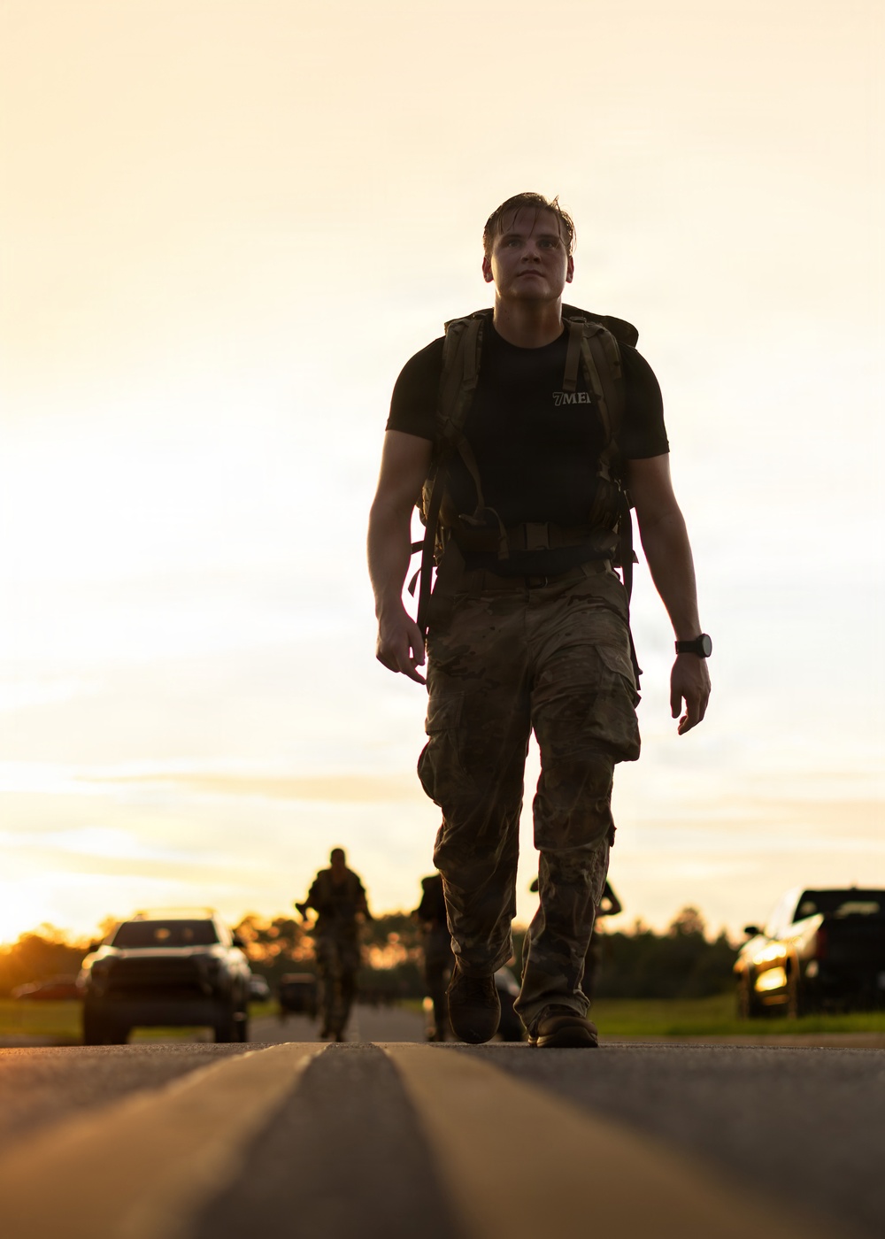 A young male in military-style attire walks purposefully down a road toward the camera during golden hour lighting conditions. He is wearing a black t-shirt with partial text '7ME[D]', camouflage cargo pants, a tactical backpack, and a watch. Additional figures and vehicles are visible in the background, suggesting a group activity such as a ruck march or physical training event.