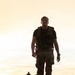A young male in military-style attire walks purposefully down a road toward the camera during golden hour lighting conditions. He is wearing a black t-shirt with partial text '7ME[D]', camouflage cargo pants, a tactical backpack, and a watch. Additional figures and vehicles are visible in the background, suggesting a group activity such as a ruck march or physical training event.