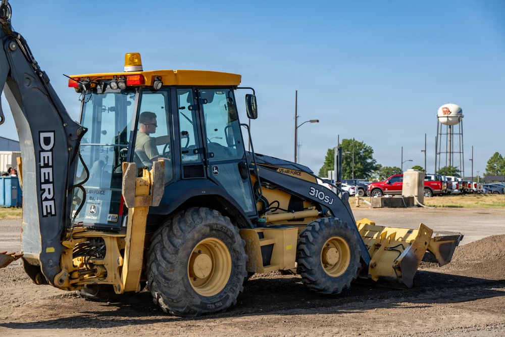 A John Deere 310SJ backhoe loader marked 'US Air Force' is being operated at what appears to be a military base construction or earthmoving site. A single operator in military-style clothing is visible inside the enclosed cab. The machine is positioned on disturbed dirt ground with a parking lot, water tower, and trees visible in the background.