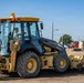 A John Deere 310SJ backhoe loader marked 'US Air Force' is being operated at what appears to be a military base construction or earthmoving site. A single operator in military-style clothing is visible inside the enclosed cab. The machine is positioned on disturbed dirt ground with a parking lot, water tower, and trees visible in the background.