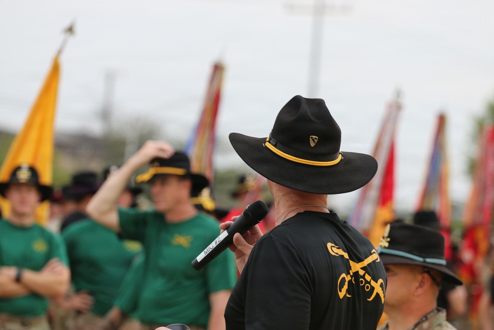 A military ceremony or formation event featuring personnel wearing distinctive black and olive/green cavalry-style hats. The foreground figure, seen from behind, is speaking into a microphone while wearing a black shirt with crossed sabers insignia and a black cavalry hat with gold cord — consistent with U.S. Army 1st Cavalry Division. Multiple unit guidons and flags are visible in the background.