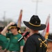 A military ceremony or formation event featuring personnel wearing distinctive black and olive/green cavalry-style hats. The foreground figure, seen from behind, is speaking into a microphone while wearing a black shirt with crossed sabers insignia and a black cavalry hat with gold cord — consistent with U.S. Army 1st Cavalry Division. Multiple unit guidons and flags are visible in the background.