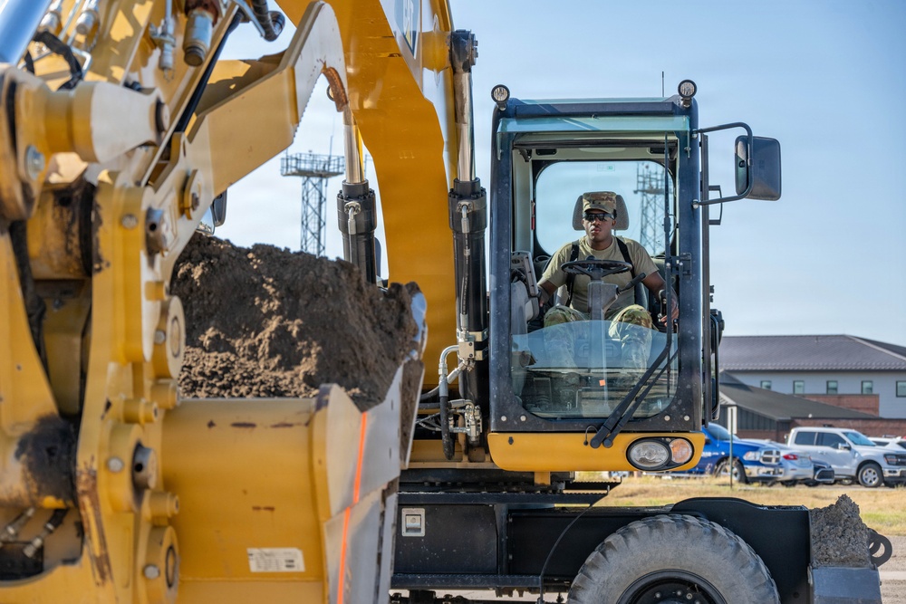 A military service member in uniform and cap is operating a large yellow CAT (Caterpillar) wheeled excavator, with the bucket visibly loaded with dirt or soil. The scene appears to take place on or near a military installation, evidenced by the operator's military attire and institutional buildings visible in the background. The photograph is taken from a low, close-up angle emphasizing the scale of the heavy equipment.