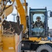 A military service member in uniform and cap is operating a large yellow CAT (Caterpillar) wheeled excavator, with the bucket visibly loaded with dirt or soil. The scene appears to take place on or near a military installation, evidenced by the operator's military attire and institutional buildings visible in the background. The photograph is taken from a low, close-up angle emphasizing the scale of the heavy equipment.