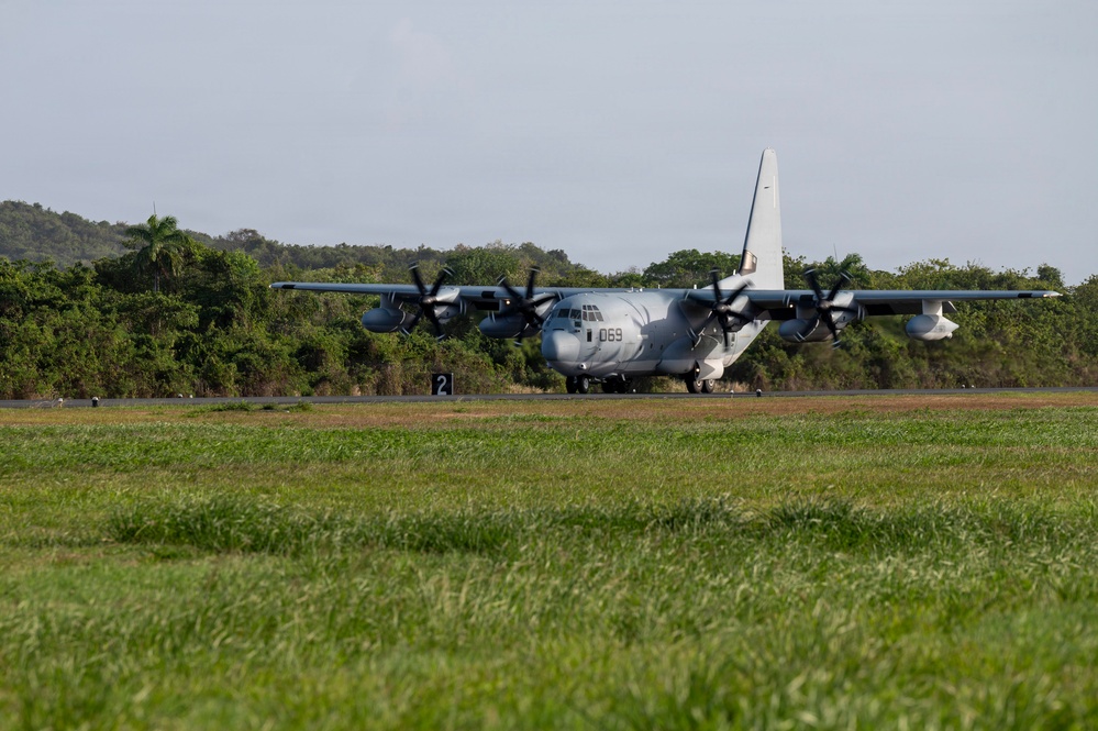 A military turboprop transport aircraft, identifiable as a C-130 Hercules variant with tail number '069', is taxiing or positioned on a runway. The aircraft is painted in a light gray military scheme and features four turboprop engines. The setting appears to be a tropical or subtropical airfield, surrounded by dense green vegetation and hills.