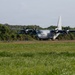 A military turboprop transport aircraft, identifiable as a C-130 Hercules variant with tail number '069', is taxiing or positioned on a runway. The aircraft is painted in a light gray military scheme and features four turboprop engines. The setting appears to be a tropical or subtropical airfield, surrounded by dense green vegetation and hills.