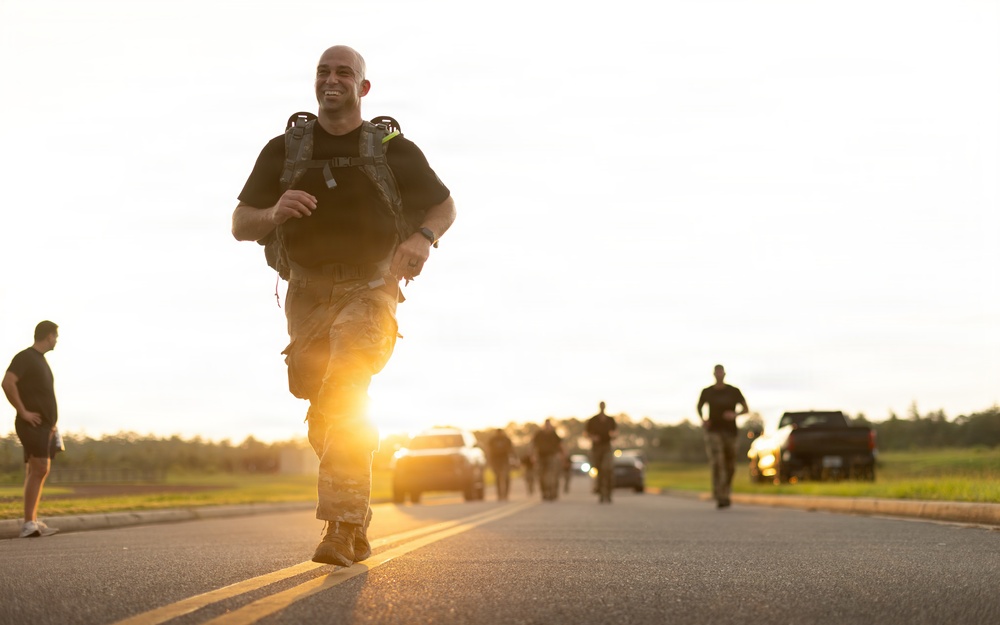 A smiling bald man in military camouflage pants and a black t-shirt runs along a road while carrying a loaded tactical backpack, likely participating in a military ruck march or fitness event. The image is taken at golden hour with a bright sun flare in the background, illuminating a line of other participants and vehicles behind him. The overall atmosphere suggests a structured military physical training exercise.