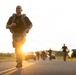 A smiling bald man in military camouflage pants and a black t-shirt runs along a road while carrying a loaded tactical backpack, likely participating in a military ruck march or fitness event. The image is taken at golden hour with a bright sun flare in the background, illuminating a line of other participants and vehicles behind him. The overall atmosphere suggests a structured military physical training exercise.