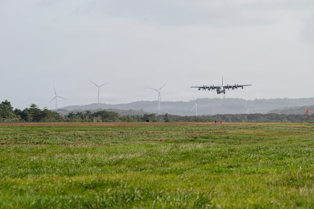 A military turboprop aircraft, consistent with a C-130 Hercules, is captured on final approach or low-altitude flight over a grassy airfield. The background features multiple wind turbines, rolling tree-covered hills, and overcast skies. The scene appears to be a military or joint-use airfield in a tropical or subtropical region.