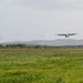 A military turboprop aircraft, consistent with a C-130 Hercules, is captured on final approach or low-altitude flight over a grassy airfield. The background features multiple wind turbines, rolling tree-covered hills, and overcast skies. The scene appears to be a military or joint-use airfield in a tropical or subtropical region.