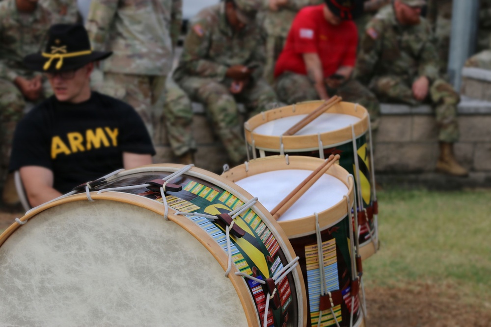 The image shows a U.S. Army military drum line or band setting, with decorated snare/tenor drums prominently displayed in the foreground. A soldier wearing an 'ARMY' t-shirt and a black cavalry hat with crossed sabers insignia is visible in the background. Multiple soldiers in camouflage uniforms are seated in the background, suggesting a military ceremony or event.