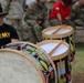 The image shows a U.S. Army military drum line or band setting, with decorated snare/tenor drums prominently displayed in the foreground. A soldier wearing an 'ARMY' t-shirt and a black cavalry hat with crossed sabers insignia is visible in the background. Multiple soldiers in camouflage uniforms are seated in the background, suggesting a military ceremony or event.