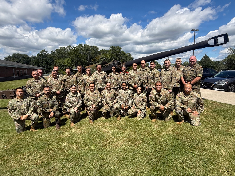 A group of approximately 25 military personnel in U.S. Army OCP (Operational Camouflage Pattern) uniforms pose for a formal group photograph on a grass lawn. They are arranged in two rows — a kneeling front row and a standing back row — in front of what appears to be a static display military tank or artillery piece. The setting appears to be a military installation or armory, likely during a training event or unit gathering.