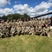 A group of approximately 25 military personnel in U.S. Army OCP (Operational Camouflage Pattern) uniforms pose for a formal group photograph on a grass lawn. They are arranged in two rows — a kneeling front row and a standing back row — in front of what appears to be a static display military tank or artillery piece. The setting appears to be a military installation or armory, likely during a training event or unit gathering.