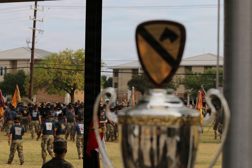 The image shows a military formation ceremony viewed through a window, with a trophy in the foreground bearing the 1st Cavalry Division patch (black horse head on gold shield). Soldiers in both physical training shirts and Army Combat Uniforms are assembled on a parade field, with unit guidons and flags visible. The scene appears to be a military competition award ceremony or unit formation at what is likely Fort Cavazos (formerly Fort Hood), Texas.