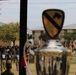 The image shows a military formation ceremony viewed through a window, with a trophy in the foreground bearing the 1st Cavalry Division patch (black horse head on gold shield). Soldiers in both physical training shirts and Army Combat Uniforms are assembled on a parade field, with unit guidons and flags visible. The scene appears to be a military competition award ceremony or unit formation at what is likely Fort Cavazos (formerly Fort Hood), Texas.