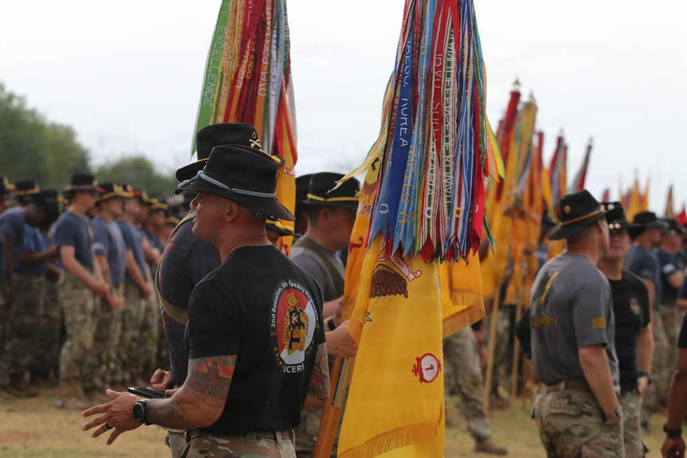 A U.S. Army military formation ceremony featuring soldiers in physical training attire and campaign hats carrying unit guidons and colors adorned with numerous campaign and award streamers. The prominent yellow flags with gold fringe are consistent with U.S. Army cavalry unit colors. The event appears to be a formal regimental or brigade-level ceremony, likely at a U.S. Army installation.