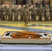 A large rectangular sheet cake decorated with an orange and black tiger mascot design is displayed in the foreground at what appears to be a military ceremony. In the background, numerous soldiers in camouflage uniforms are assembled in formation on a field featuring a large yellow and black chevron or unit insignia painted on the ground. The setting suggests a unit anniversary, activation, or celebratory military event.