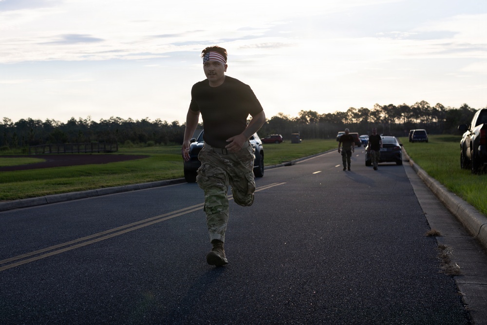 A person in military camouflage pants and a black t-shirt is running along a paved road, wearing an American flag headband. The setting appears to be a military base or installation during early morning or evening hours based on the low-angle lighting. Additional personnel and parked vehicles are visible in the background.