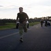 A person in military camouflage pants and a black t-shirt is running along a paved road, wearing an American flag headband. The setting appears to be a military base or installation during early morning or evening hours based on the low-angle lighting. Additional personnel and parked vehicles are visible in the background.
