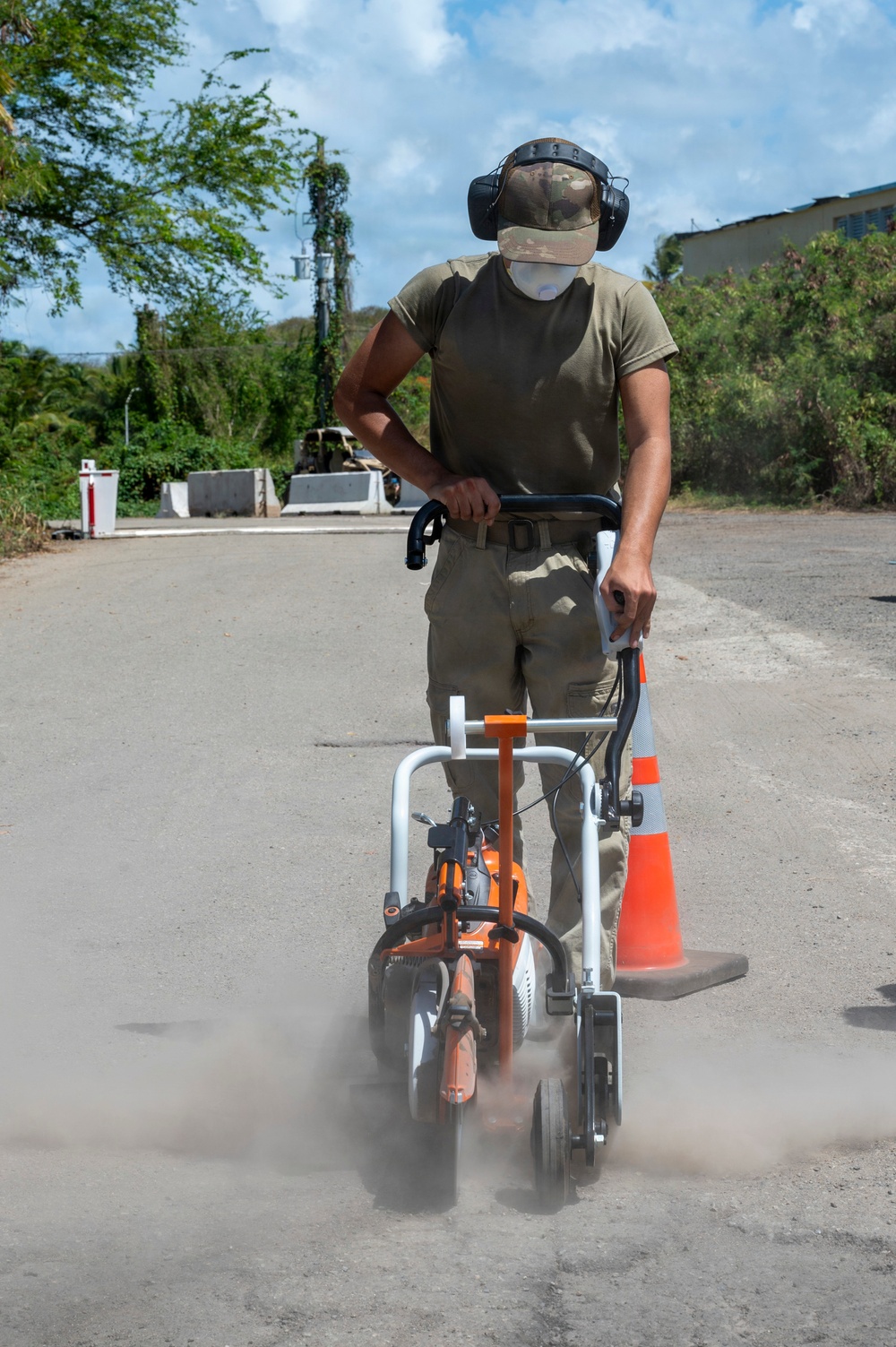 A person in military-style clothing operates a walk-behind concrete cutting or grinding machine on a paved road surface, generating visible dust. The operator is wearing appropriate personal protective equipment including hearing protection, a dust mask, and a camouflage cap. An orange traffic cone is positioned nearby, and the setting appears to be an outdoor road or base facility surrounded by tropical vegetation.