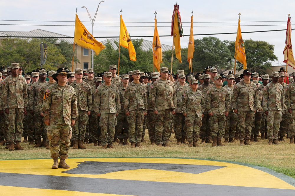 A large formation of U.S. Army soldiers in OCP (Operational Camouflage Pattern) uniforms stands at attention during a formal military ceremony. Multiple unit guidons (yellow/gold flags) are visible being carried by flag bearers throughout the formation. The ceremony appears to be taking place on a parade field featuring a large yellow and black unit insignia painted on the ground.