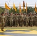 A large formation of U.S. Army soldiers in OCP (Operational Camouflage Pattern) uniforms stands at attention during a formal military ceremony. Multiple unit guidons (yellow/gold flags) are visible being carried by flag bearers throughout the formation. The ceremony appears to be taking place on a parade field featuring a large yellow and black unit insignia painted on the ground.