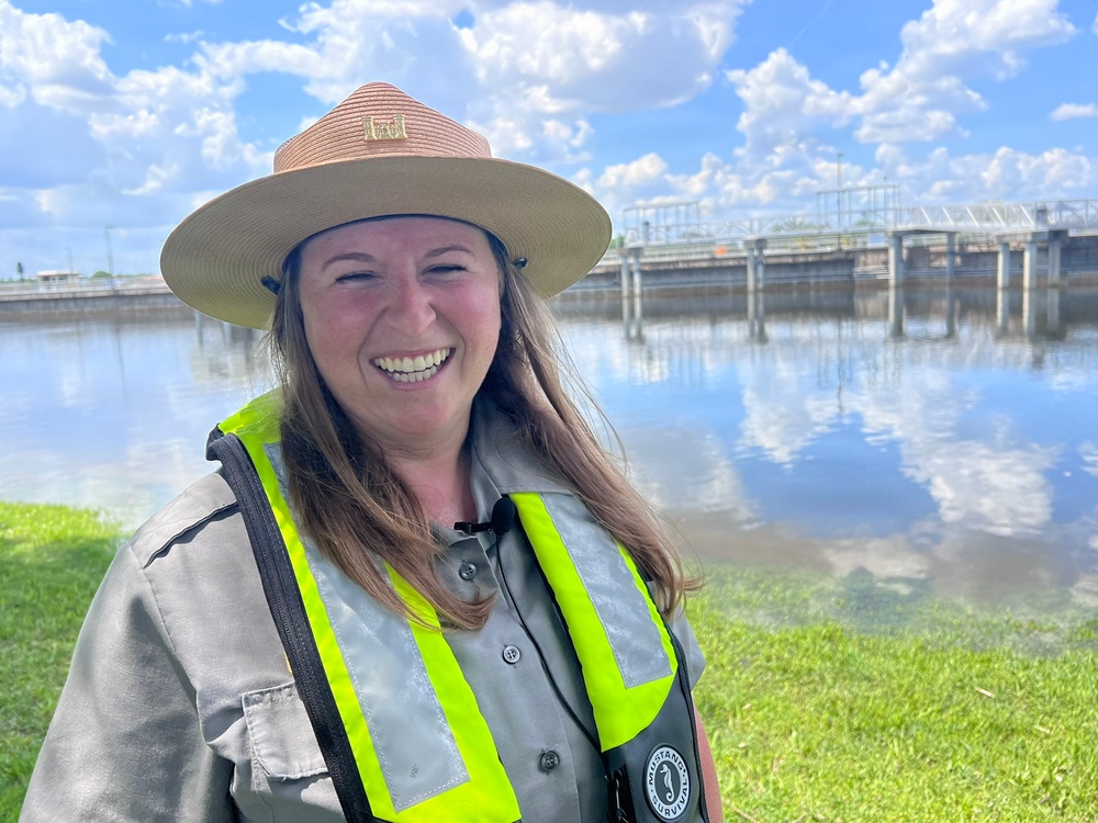 A woman wearing a ranger-style flat-brimmed hat and a high-visibility safety vest over a gray uniform shirt stands smiling in front of a water control structure or lock. She appears to be a park ranger, water management official, or safety officer working near a waterway. The background shows a calm body of water reflecting clouds and a concrete water control gate or dam structure.