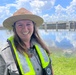 A woman wearing a ranger-style flat-brimmed hat and a high-visibility safety vest over a gray uniform shirt stands smiling in front of a water control structure or lock. She appears to be a park ranger, water management official, or safety officer working near a waterway. The background shows a calm body of water reflecting clouds and a concrete water control gate or dam structure.