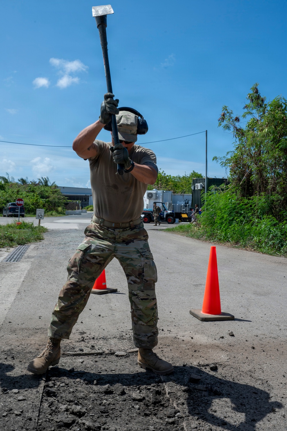 A military service member in MultiCam/OCP camouflage uniform is using a sledgehammer to break up road pavement on a paved road. The work area is marked with orange traffic cones, and additional personnel and equipment are visible in the background. The setting appears to be a tropical or subtropical location based on the lush vegetation and blue sky.