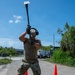 A military service member in MultiCam/OCP camouflage uniform is using a sledgehammer to break up road pavement on a paved road. The work area is marked with orange traffic cones, and additional personnel and equipment are visible in the background. The setting appears to be a tropical or subtropical location based on the lush vegetation and blue sky.